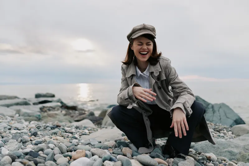 Happy young woman wearing a coat and hat laughs joyfully while sitting on a rocky beach during cloudy weather. Concept of happiness, nature, and casual outdoor lifestyle.