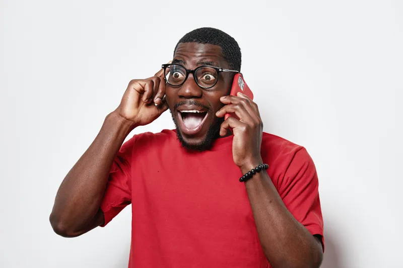 African American man with dark skin and glasses wearing red shirt talking on phone with excited expression and hand near ear on white background. Communication, emotion and technology.