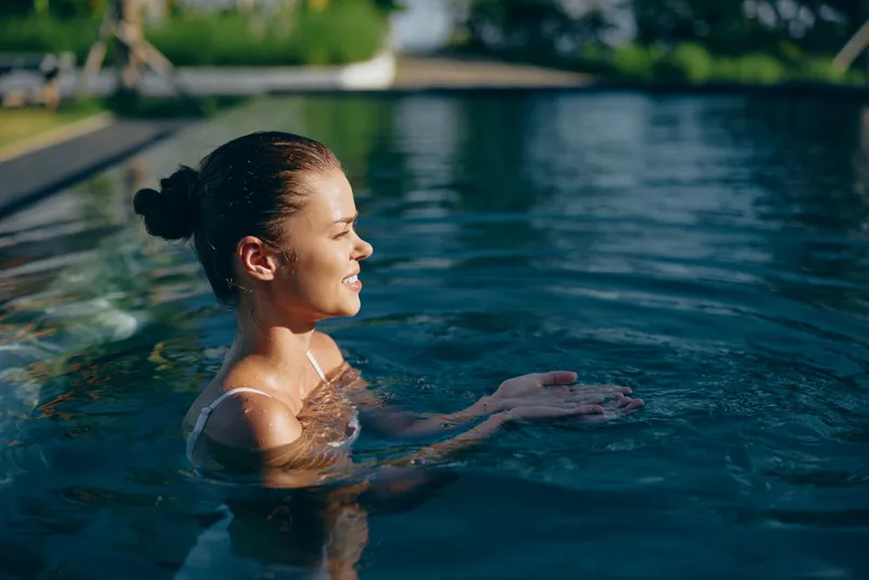 Young woman with brunette hair enjoys a peaceful moment swimming in an outdoor pool under soft sunlight with relaxed expression and calm water around her.