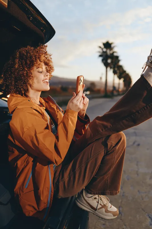 Woman lifestyle outdoors, curly hair smiling, holding phone for casual selfie, warm sunset light, relaxed posture, urban street background, bright evening scene