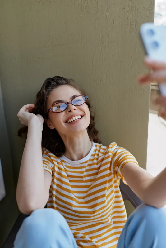 Smiling woman with striped yellow shirt taking a selfie in a home setting, casual lifestyle, relaxed mood, natural daylight, soft indoor lighting, candid portrait.