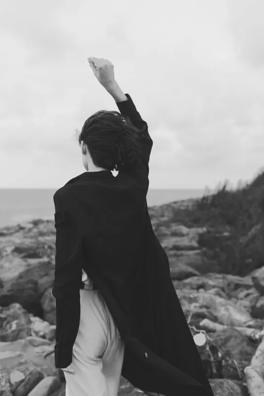 Silhouette on a rocky coast, cape fluttering, fist raised toward the windy horizon, high contrast black and white, symbolizing freedom, power, and resilience