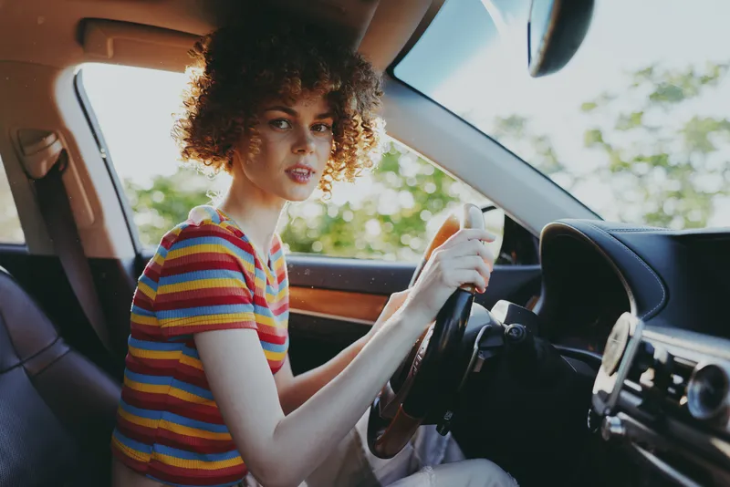 A woman driver sits behind the wheel in a striped tee, capturing a casual lifestyle moment with a retro vintage color palette in a sunlit car interior.