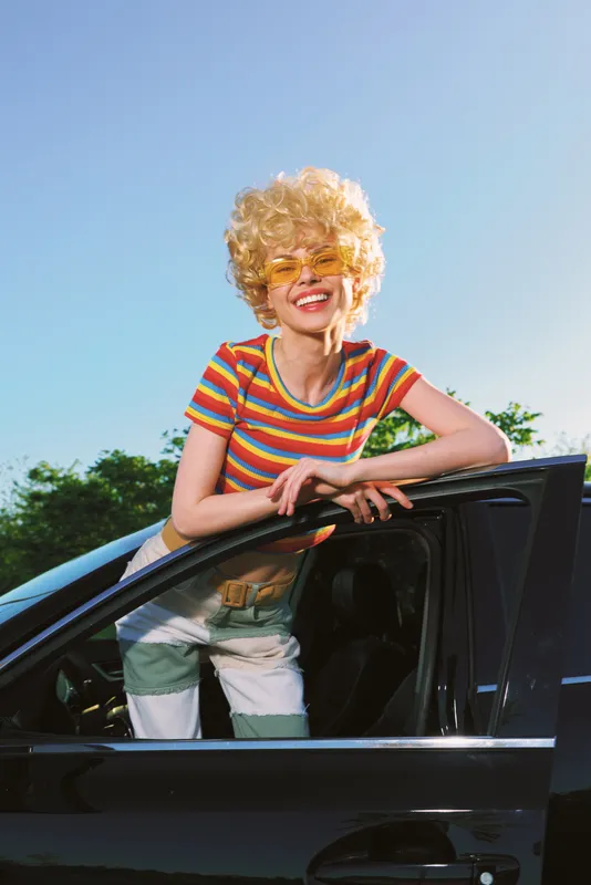 Woman with retro vibe leaning from car window, striped shirt and curly blonde wig, sunny outdoor scene, casual lifestyle, bright colors, playful mood, candid fashion moment.