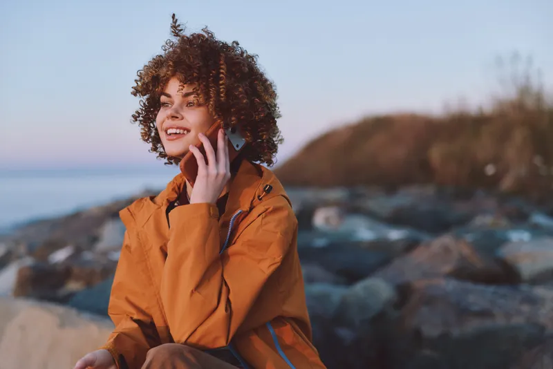 Smiling woman with curly hair in an orange jacket uses a smartphone by a rocky beach, sea waves blurred. Outdoor lifestyle travel mood with warm daylight, communication fresh air.