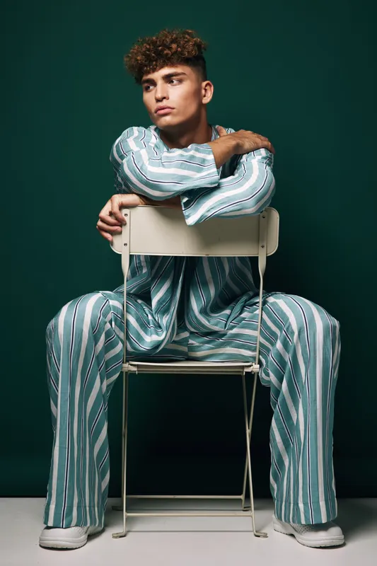Young man sitting on chair backwards posing in striped casual outfit and white sneakers against dark green background looking thoughtful and stylish in portrait studio environment.