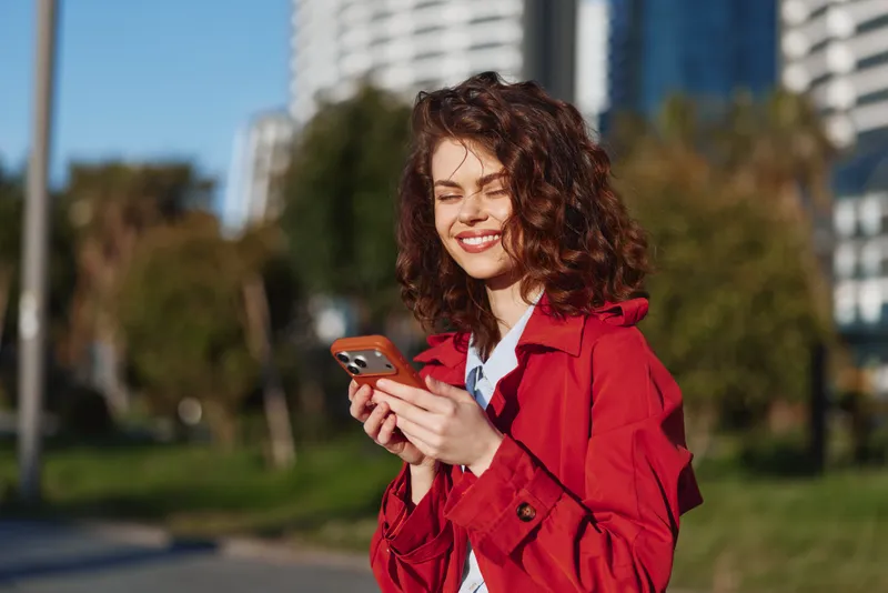 Smiling woman with curly hair wearing a red jacket checks her smartphone in a sunny park. Casual lifestyle, outdoor moment, bright mood, candid urban greenery and vitality.