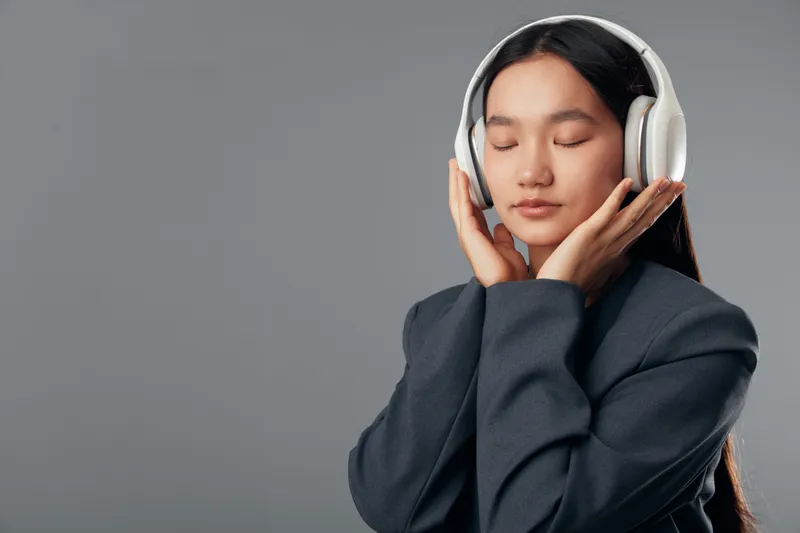 Young woman with closed eyes wearing headphones, enjoying music and relaxation in studio environment. Calm expression, long hair, casual clothing on gray background.