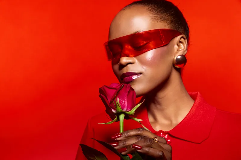 Stylish African woman with dark skin wearing red sunglasses and red outfit holds a red rose near her face on a vibrant red background. Concept of fashion, beauty, and elegance.