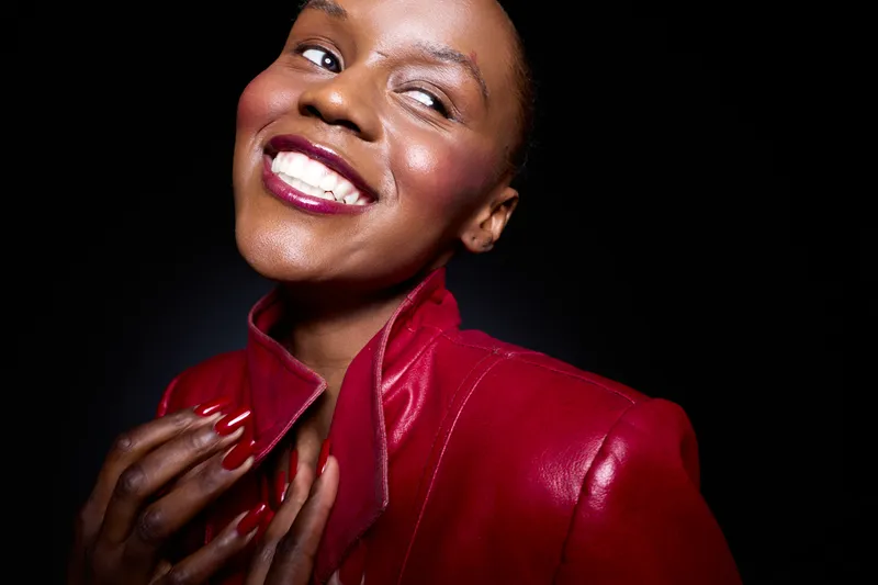 Smiling dark skinned African American woman in red leather jacket poses confidently against black background. Concept of beauty, fashion, elegance, and self-expression with joyful mood.