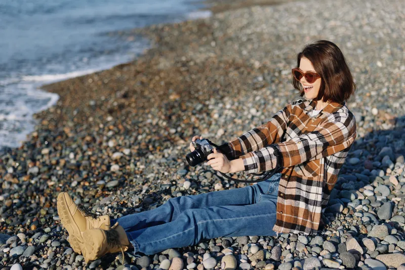 Woman on a beach wearing a plaid shirt and blue jeans sits on pebbles by the water, capturing candid photography with a handheld camera on a sunny outdoor day.