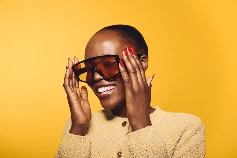 Smiling black woman with dark skin wearing oversized sunglasses and beige sweater poses against yellow background. Concept of fashion, style, happiness, and confident female model with nails.