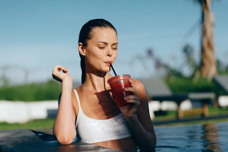 Woman in a white swimsuit drinks a refreshing smoothie by the pool, a sunlit outdoor moment, relaxed posture, warm breeze, natural colors, lifestyle scene, summertime