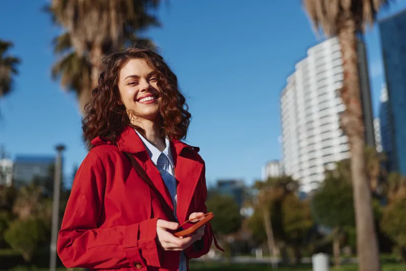 A young woman with curly hair wears a red jacket and white shirt in an urban park, smiling while holding a phone against a bright sunny day with palm trees and tall buildings nearby.