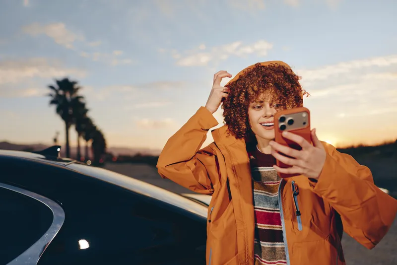 Woman lifestyle portrait outdoors at sunset beside a car, holding a smartphone for a selfie, curly hair loose, smiling in warm golden light, casual jacket, evening travel relaxed today