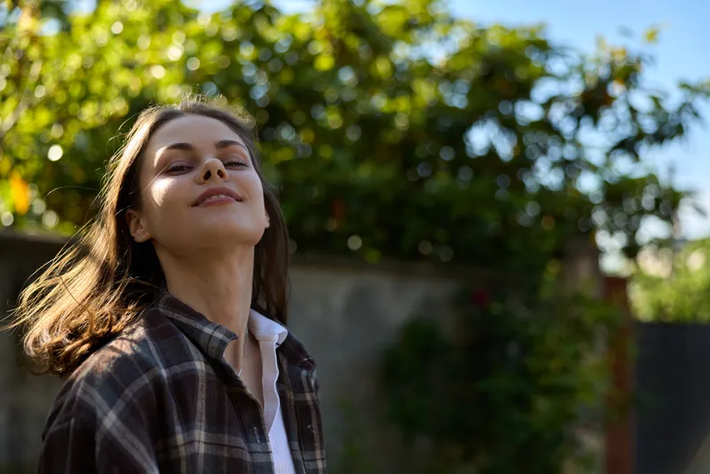 Young woman with brown hair enjoys a sunny moment outdoors, eyes closed and head tilted back, wearing a plaid shirt and white tee, radiating calm and carefree mood under warm garden light and a soft