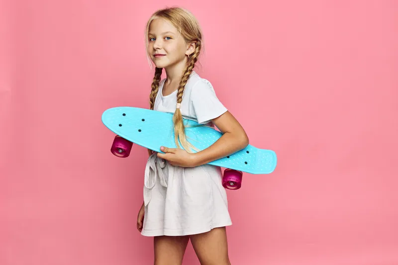 Young girl with blonde braids holds a blue skateboard on pink background. She wears a white dress and smiles confidently.