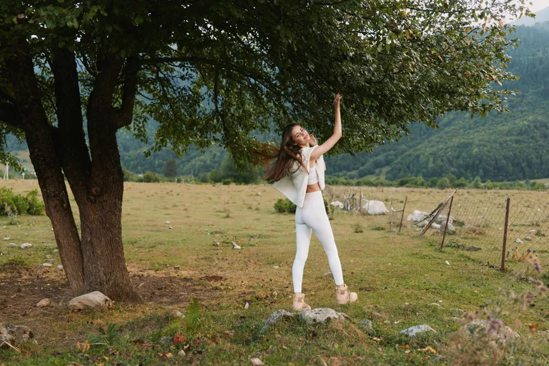 A woman dances near a large tree in an open field, wearing white clothing, smiling under sunny skies. A lifestyle moment capturing joy and freedom in nature. Bright countryside.