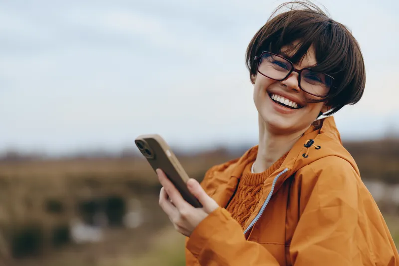 Woman lifestyle outdoors, smiling with glasses, orange jacket, holding smartphone, laughing in wind, blurred countryside background, casual travel portrait, daylight scenic horizon, closeup