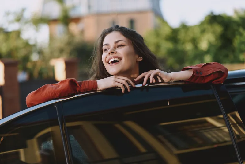 A young woman leans from a car window, laughing with carefree joy during a casual city drive, capturing a candid moment of happiness and freedom, Her red jacket adds energy to the frame