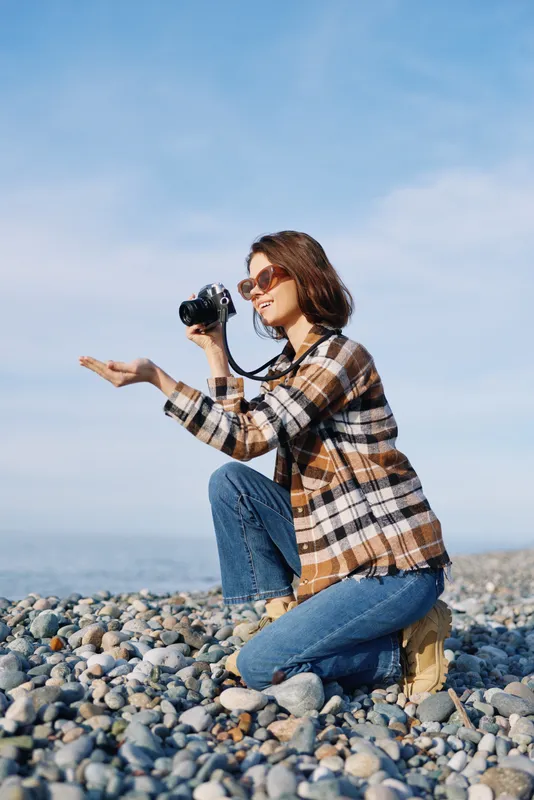 Woman photographer kneels on a pebble beach, plaid shirt and jeans, sunglasses on, camera raised to capture coastal scenery under a clear blue sky.