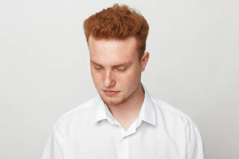 Young man with red hair and fair skin looks down wearing a white shirt on a plain light background. Concept of introspection, contemplation, and calm mood in portrait photography.