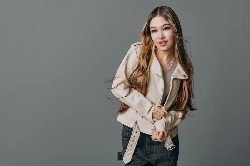 Young woman with long hair wearing a beige jacket and dark jeans posing on gray background. Female model looking down with hands adjusting jacket belt in casual style.