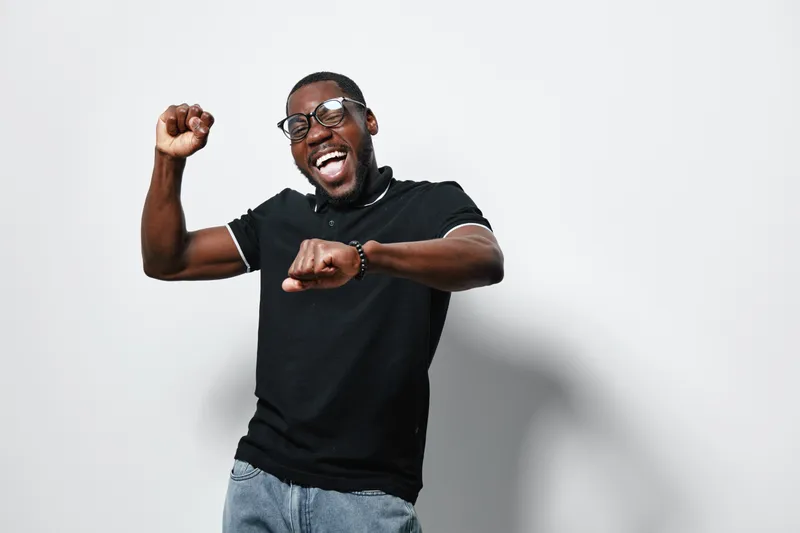 African American man with dark skin wearing glasses and black polo shirt dancing happily with raised fists on white background. Casual style and joyful expression.