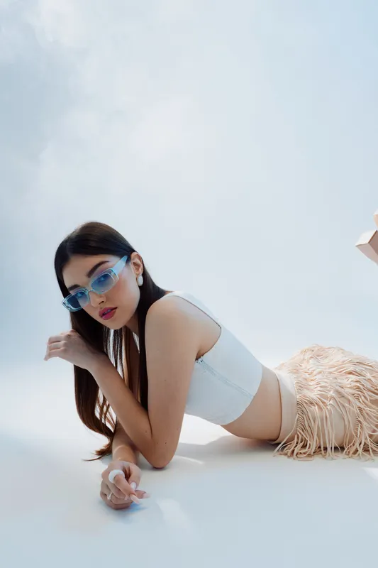 Young woman in stylish blue sunglasses and white crop top poses lying down on a white floor. Concept of woman beauty fashion with modern accessories and casual style.