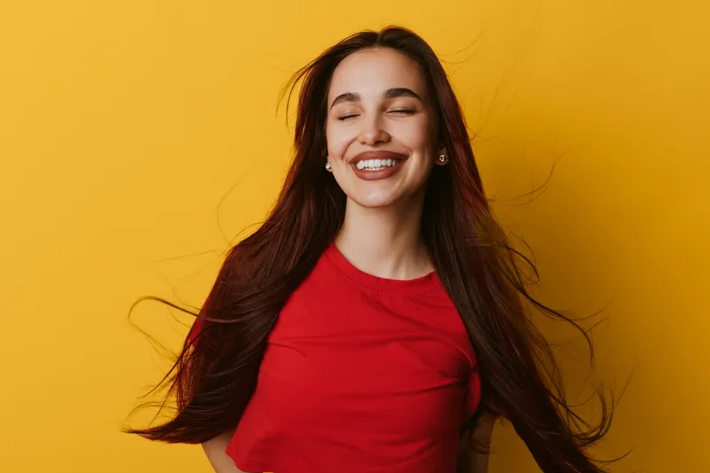 Young woman with long hair smiling with closed eyes against yellow background. Female model wearing red shirt expressing happiness and joy with natural beauty and relaxed pose.
