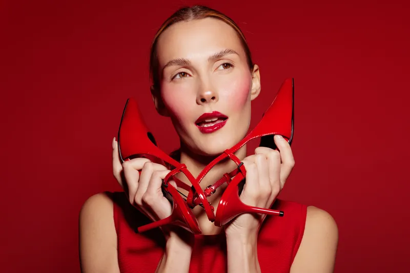 Young woman in red dress holds red high heels near face on red background. Fashion concept with matching colors, beauty, style, and confident female model in studio lighting.