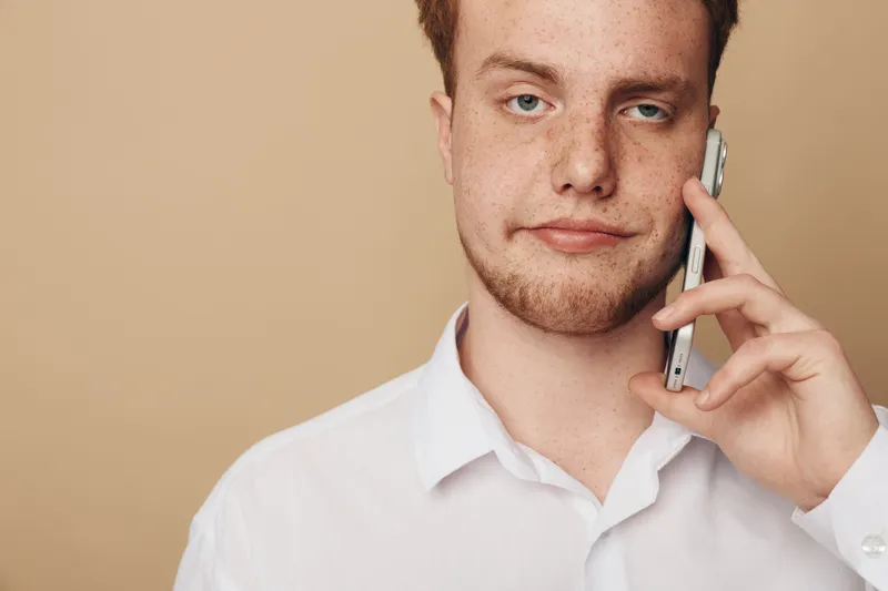 Young man with red hair and freckles talks on smartphone, wearing white shirt against beige background. Concept of communication, technology, and casual lifestyle.