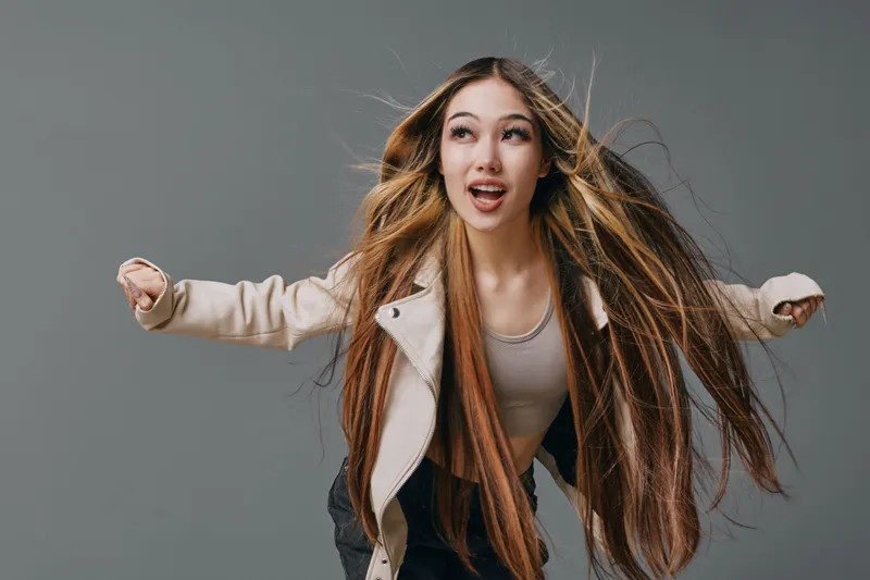Young woman with long hair wearing casual jacket and top posing energetically on grey background with dynamic expression and movement of hair in motion.