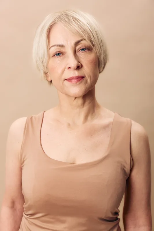 Portrait of confident mature woman with short blonde hair wearing beige tank top against neutral background, showing natural beauty and calm expression in soft lighting.