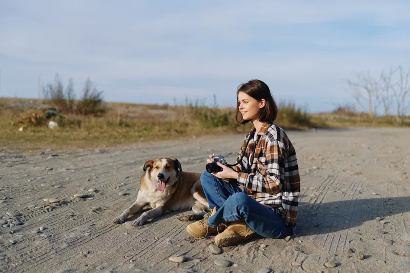 Woman, dog, outdoors, desert landscape with clear sky, sitting on sandy ground and holding a camera, enjoying companionship, nature, and a relaxed casual moment