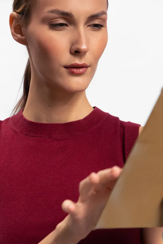 Young woman in red sweater closely examines a brown paper sheet, focused expression. Studio lighting highlights natural skin tone and details. Concept of concentration, study, and creativity.