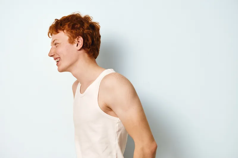 Young man with red hair wearing sleeveless shirt smiling and laughing in casual pose isolated on white background. Profile portrait of cheerful teenager with fair skin in studio.