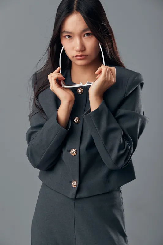 Young woman with long dark hair and gray suit holding sunglasses close to her face in studio. Fashion model posing seriously with modern outfit on neutral background.