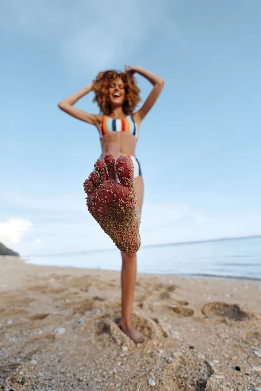 woman beach summer happiness swimsuit curly pose sunlight ocean, smiling model stands on sandy shoreline with arms on head, blue sky and waves in background, vacation leisure relaxation, natural