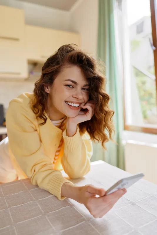 A cheerful woman sits at a bright home bedroom, smiling into a smartphone for a casual lifestyle moment, cozy, intimate mood, candid self portrait in a sunny space