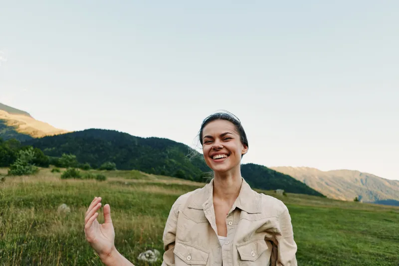 A laughing young woman stands in a sunny field with a mountain backdrop, arms relaxed, looking toward the camera, radiating warmth and carefree energy in daylight outdoors.
