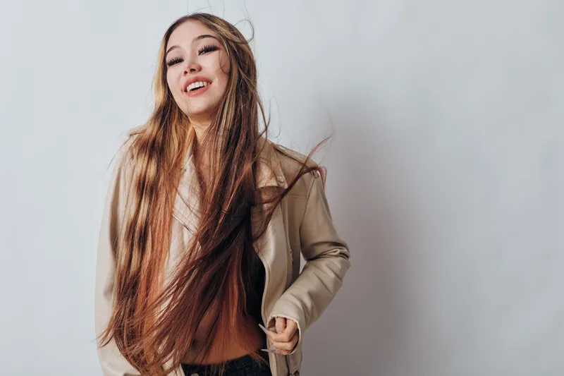 Young woman with long hair smiling and wearing light jacket against white background. Casual style, fashion portrait, confident person enjoying moment indoors.