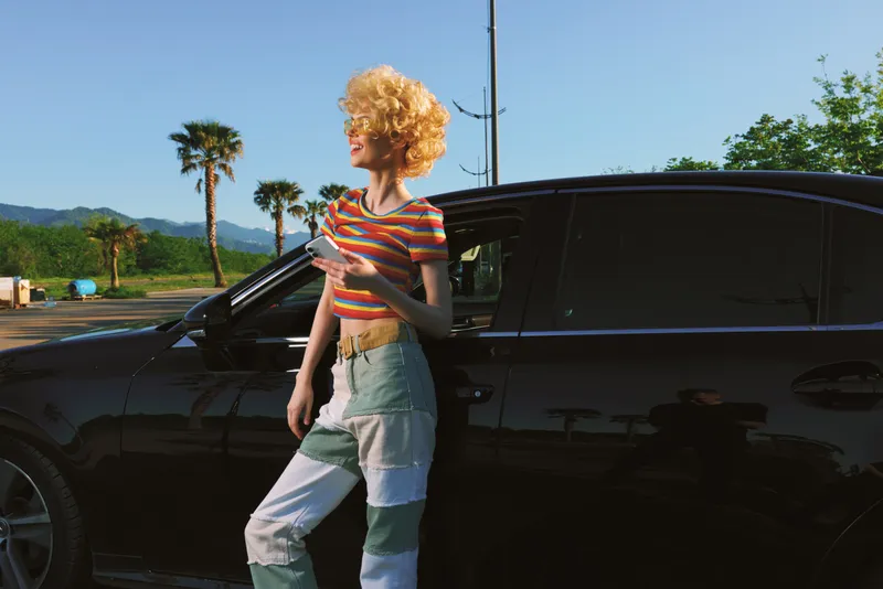 A stylish young woman with curly blonde hair stands beside a dark car in a bright lot, wearing a striped top and patchwork jeans, radiating a retro, carefree road trip vibe.