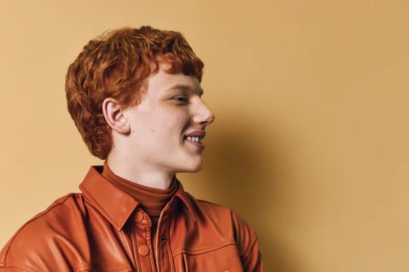 Young man with red hair and freckles wearing orange leather jacket smiling and looking sideways against beige background in studio portrait.