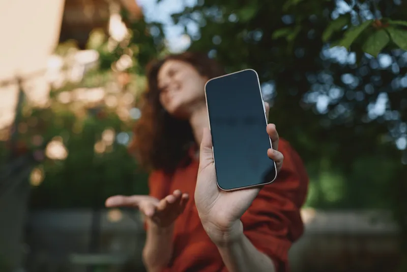 A young woman outdoors extends her arm toward the camera, showing a smartphone in focus while smiling, creating a candid lifestyle moment