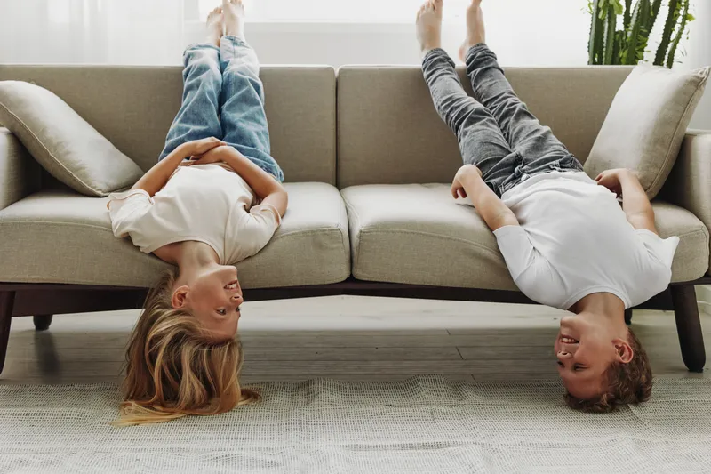 Two happy children lying upside down on a beige sofa in casual clothes laughing and looking at each other in a bright living room with natural light and a cozy atmosphere.