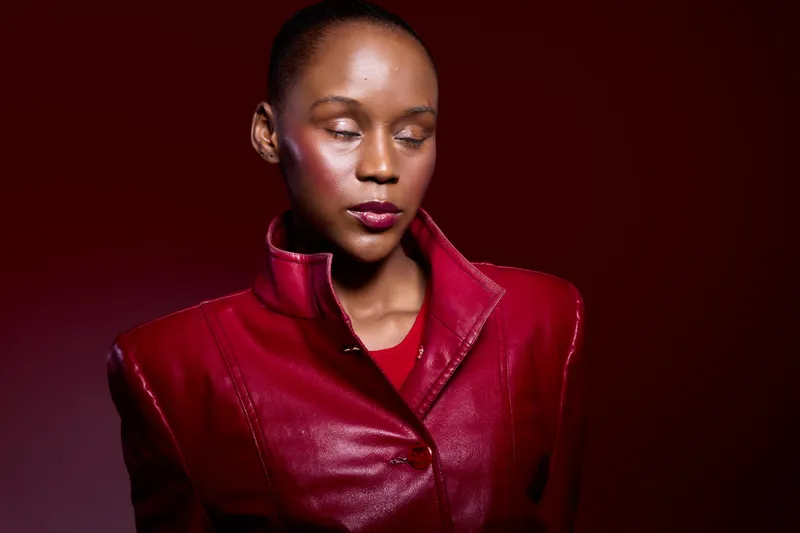 Portrait of a young black woman with dark skin wearing a red leather jacket against a dark red background. She looks down with a calm and thoughtful expression, fashion concept.