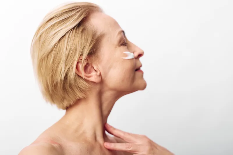 Mature woman with short blonde hair applies face cream on cheek in profile. Skincare routine concept, anti aging, self care, beauty treatment, natural skin, wellness at home.