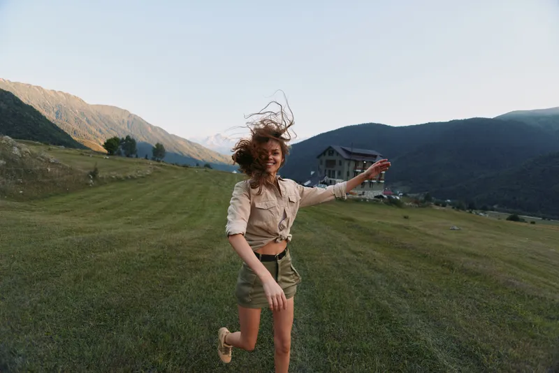 Young woman runs across a green hillside with mountains in the background, hair flying and arms outstretched, conveying carefree outdoor energy and a playful moment.