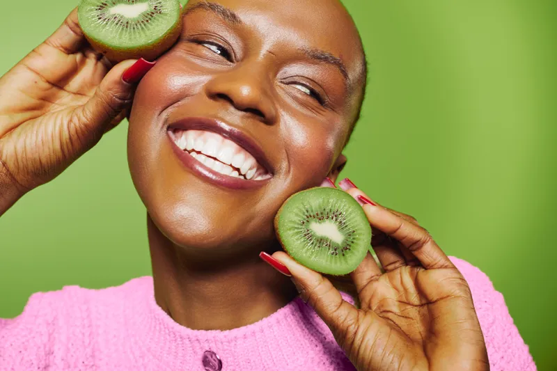 Happy black woman with dark skin holds two kiwi halves near her face smiling. Close-up portrait on green background. Concept of healthy lifestyle, natural beauty, wellness, and fresh fruit.